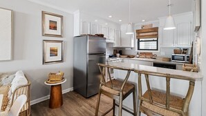 A modern kitchen with white cabinets, a stainless steel refrigerator, a wooden dining table, and a hanging light fixture. - Shell Shocked - Short, Breezy Walk to the Beach (Carolina Beach)