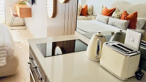 A modern kitchen with a built-in stove, a toaster, and a sink. There is a dining area with a table and chairs, and a bed with pillows in the background.
