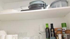 A kitchen shelf with white mugs, clear glasses, a wine glass, bottles of oil, and a spray bottle.