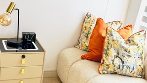 A bedroom with a beige sofa, a small wooden nightstand with a lamp, and decorative pillows.