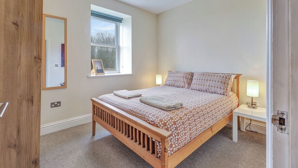 A bedroom with a wooden bed, a nightstand, a bedside lamp, a window with a view of trees, and a mirror on the wall.