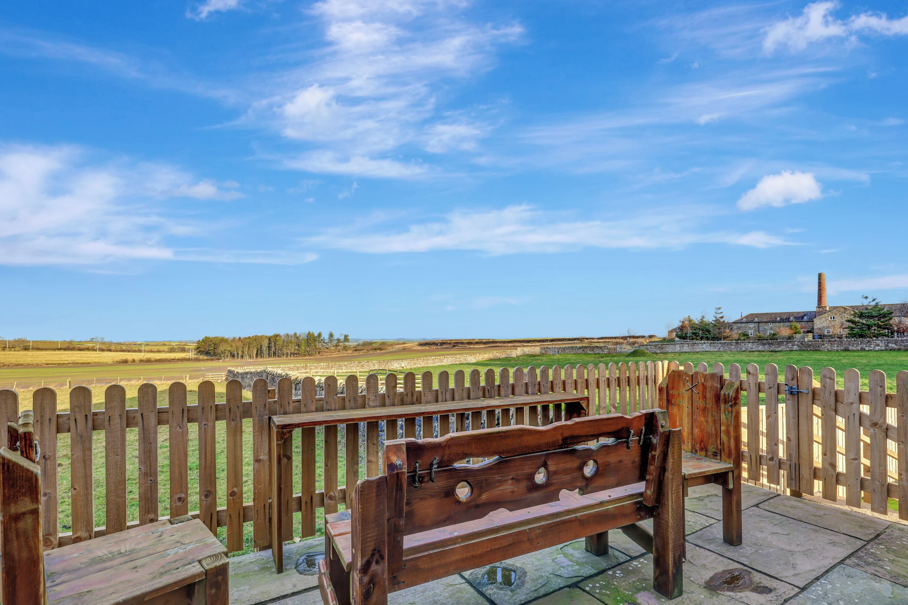 A wooden bench with a metal backrest and a metal table, set on a stone patio with a wooden fence in the background.