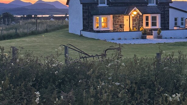 Une maison à deux étages avec une façade en pierre, une grande véranda à l’avant et une vue sur les montagnes en arrière-plan.