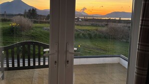 Un balcon offrant une vue sur un coucher de soleil se déclinant sur une montagne, un banc en bois et une porte vitrée.