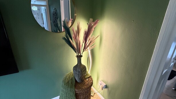 Una habitación con una pared verde, un espejo redondo, un taburete tejido y un florero con plantas secas.