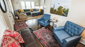 Una sala de estar con un sofá modular, un sillón azul y una mesita. Hay dos alfombras en el suelo, y un cuadro en la pared.
