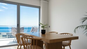 A dining area with a wooden table and chairs, a potted plant, and a view of the ocean through large sliding doors. - White Apartment (Funchal)