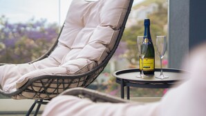 A wicker lounge chair with a cushion, a bottle of champagne, and two glasses on a small table, with a view of purple flowers and buildings in the background. - White Apartment (Funchal)