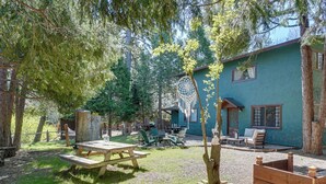 Een rustieke buitenruimte met een houten picknicktafel, een dromenvanger in een boom en een blauw gebouw met raam en deur.