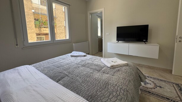 A bedroom with a bed, a TV on a white cabinet, a window, and a door.