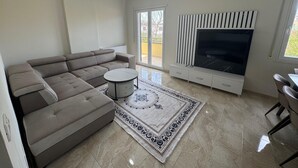 A modern living room with a sectional sofa, a round coffee table, a patterned rug, a TV on a white entertainment unit, and a sliding glass door leading to an outdoor area.