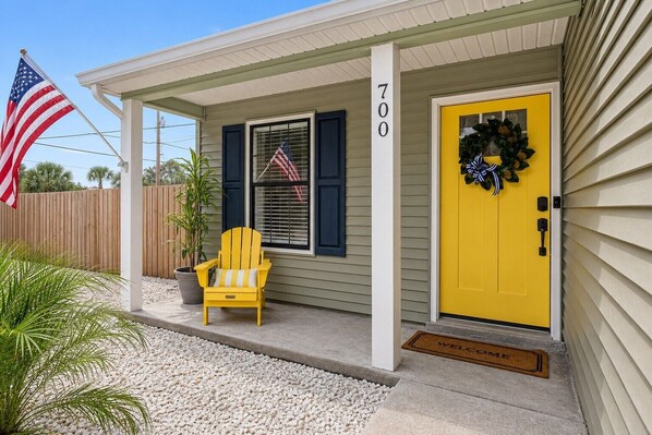 A house with a yellow door, a number 700 sign, an American flag, and a yellow Adirondack chair.