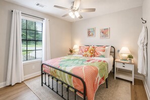 A bedroom with a bed, a bedside table, a nightstand, a ceiling fan, a window with curtains, and artwork on the wall. - Gritz & Giggles, Southern Charm Meets Belly Laughs! Close to Parris Island (Port Royal)