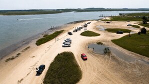 An aerial view of a parking area with vehicles, a sandy beach, and a body of water. - Gritz & Giggles, Southern Charm Meets Belly Laughs! Close to Parris Island (Port Royal)