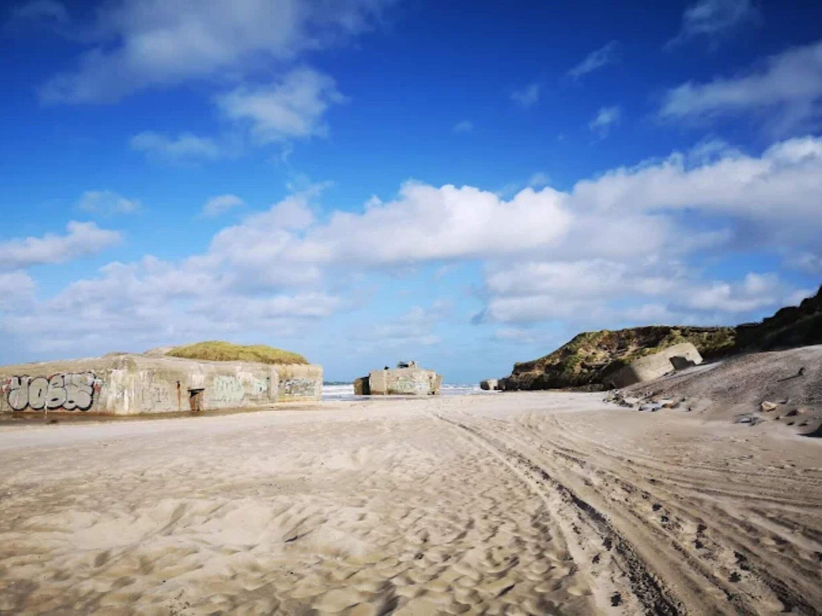 Une plage de sable avec des graffitis sur un mur, un ciel dégagé et un promontoire rocheux à l’horizon.