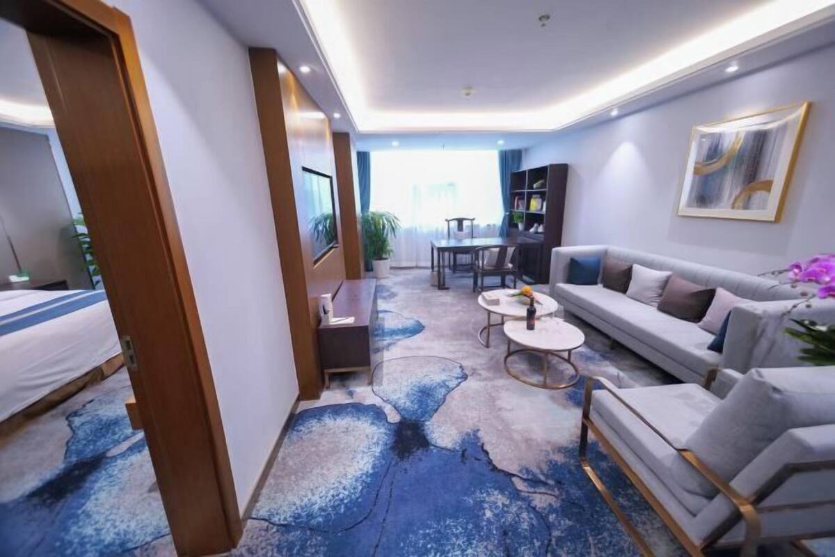 A hotel room with a bed, a sofa, a small table, a bookshelf, and a large window.