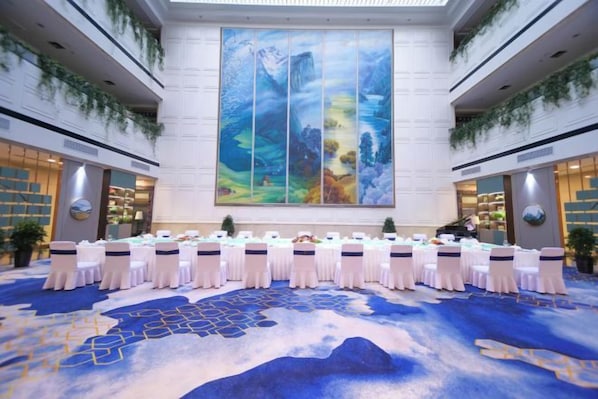Un salón de conferencias con una larga mesa dispuesta para una reunión, rodeado de sillas blancas con fajines negros, un cuadro abstracto grande en la pared y una alfombra azul con diseño de montañas.