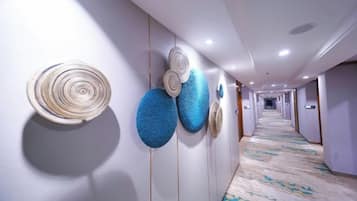 A hotel hallway with a patterned carpet, wall-mounted decorative items, and a long corridor leading to rooms.