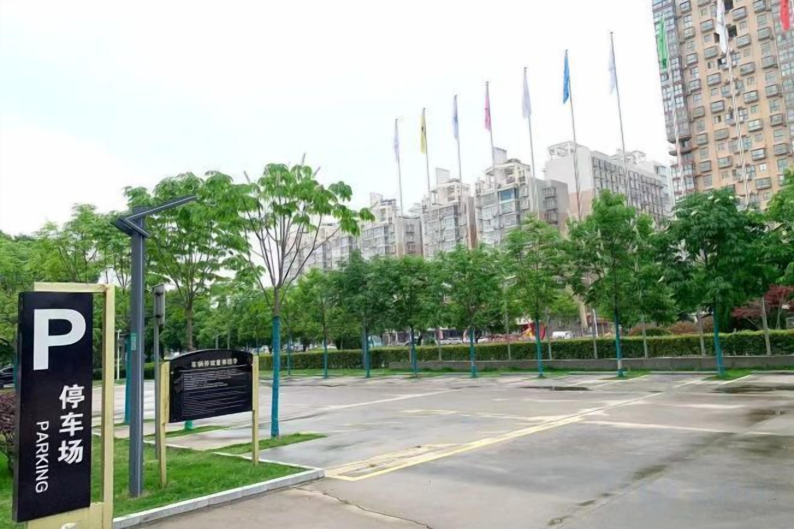 A parking lot with a sign indicating a parking area, surrounded by trees and buildings in the background.