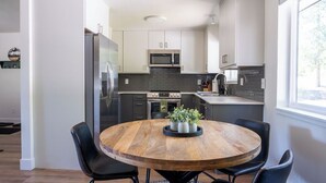 A modern kitchen with white cabinets, a stainless steel refrigerator, and a microwave. There is a round wooden table with chairs and a potted plant. - Peaceful Cabin Nestled in the Pines with a Short Walk to Lake Granby (Granby)