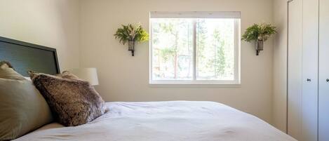A bedroom with a large bed, a bedside lamp, two wall-mounted planters, and a window with a view of trees.