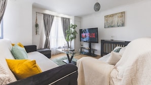 A modern living room with a sofa, armchair, and a coffee table. A television is mounted on the wall, and there is a plant on the table. - Stunning Wembley Flat | 2 Baths (Greater London)