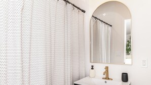 A bathroom with a white sink, a gold faucet, a black vase, and a patterned shower curtain.