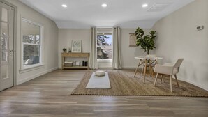 A spacious room with a wooden floor, a white sofa, a small table with chairs, a plant, and a view through a window. - Escape to comfort at Be Cozy Getaway! Located in a serene neighborhood in Euclid. (Euclid)
