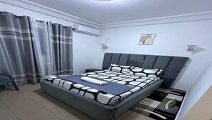 A bedroom with a bed, curtains, a nightstand, a wall-mounted air conditioner, and a small rug. - Luxury furnished studio (Douala)