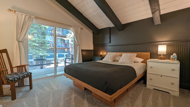 A bedroom with a large bed, a nightstand, a chair, and a view of a deck and trees outside.