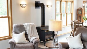 En stue med pejs, to læderlænestole, et bord med lampe, samt et spiseområde med træstole og et ur.