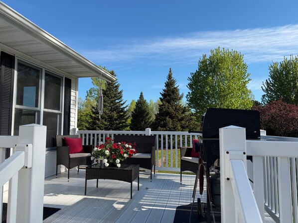 Een overdekt terras buiten met een witte balustrade, een rieten tafel met een bloemstuk en een barbecue.