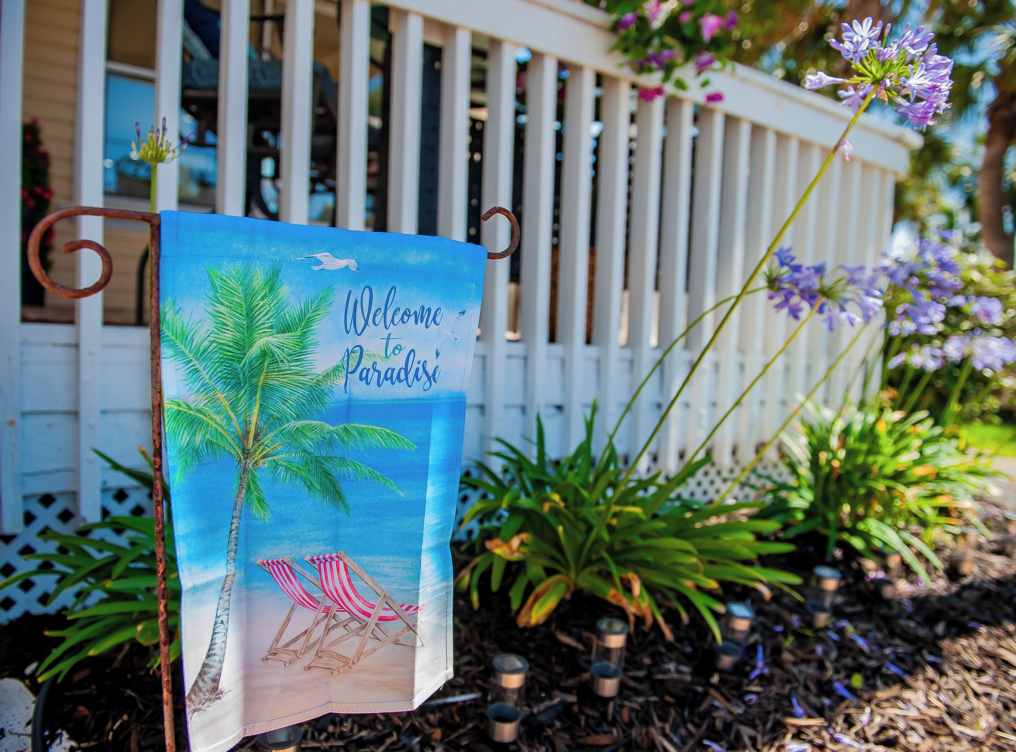 A beach-themed welcome sign with palm trees and beach chairs, placed in front of a white fence with a garden.