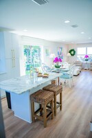 Una cocina moderna con armarios blancos, una isla de mármol y una zona de comedor con sillas blancas y una mesa preparada para cuatro.