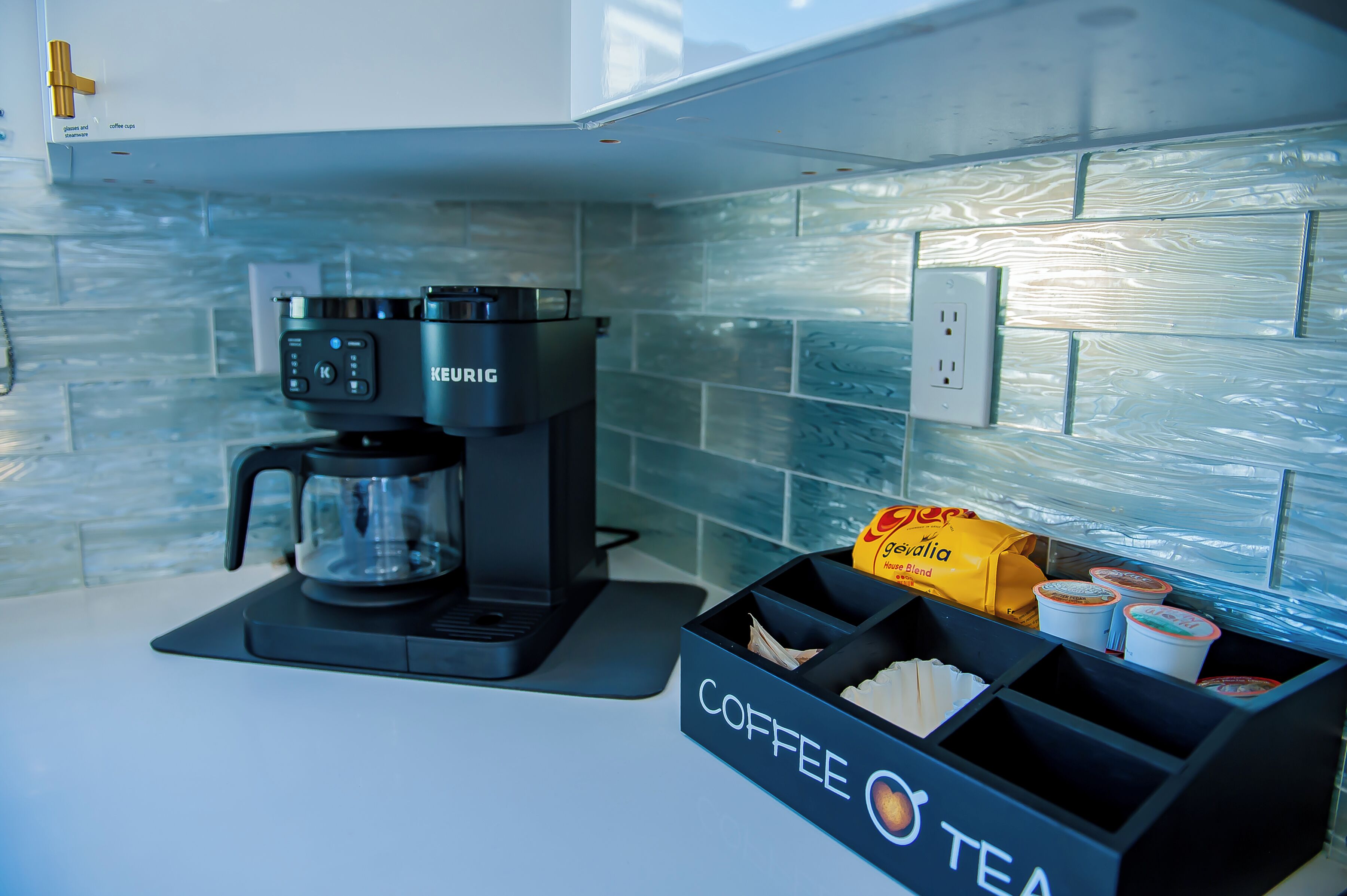 Una cafetera, una taza de café y una caja con la etiqueta 'Coffee Tea' sobre una encimera.