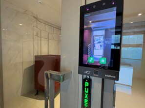 A security kiosk with a screen displaying a room layout, a metal barrier, and a sign with 'PUXE' visible.