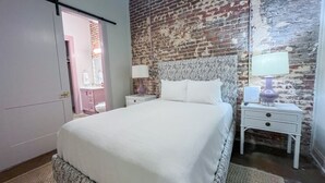 Un dormitorio con una cama grande, dos mesitas de noche, una pared con diseño de ladrillo y una puerta que da a otra habitación.