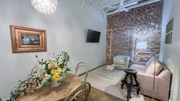 Un comedor con una mesa y sillas de madera, un centro de mesa floral y una pared de ladrillo con un televisor montado encima.