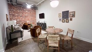 Una habitación con pared de ladrillo, una mesa redonda de madera con sillas, un sofá y un televisor montado en la pared.