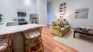 A modern kitchen with white cabinets, a microwave, and a refrigerator. There is a dining area with a white table and wicker chairs. A beige sofa is in the living room, and a coffee table is nearby. - Cane - The Flats Demopolis (Demopolis)