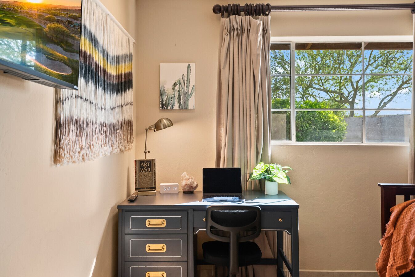 A study with a desk, a laptop, a lamp, a cactus painting, a clock, and a window with curtains.