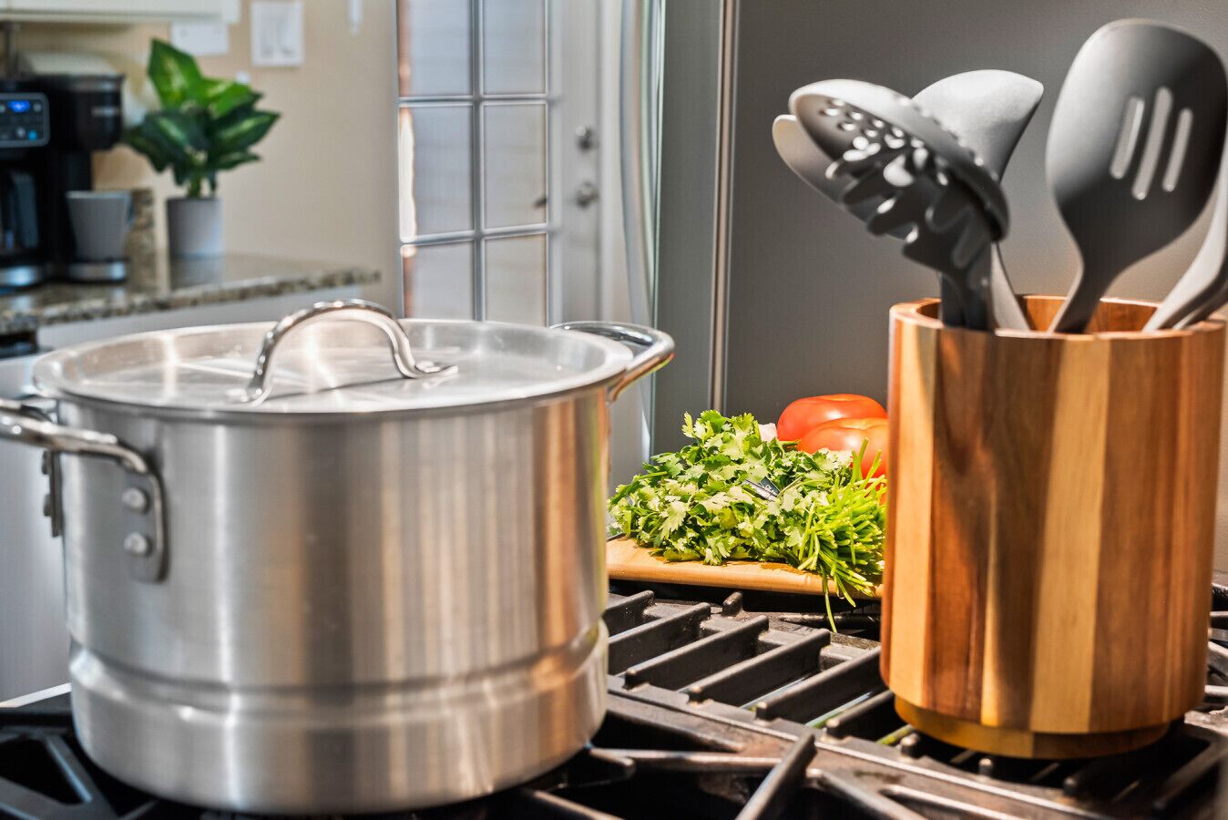 A stainless steel pot on a stove, a wooden utensil holder with spoons, and fresh vegetables on the counter.