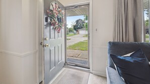 A door with a wreath, a gray door frame, a gray door, a gray wall, a gray door mat, a gray couch with a blue pillow, and a gray curtain. - Family Friendly 4 Beds/Sleeps 6 In Peaceful Westland (Westland)