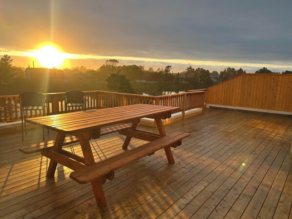 Une terrasse en bois avec des tables et des chaises pour pique-niquer, offrant une vue sur un plan d’eau et un coucher de soleil.