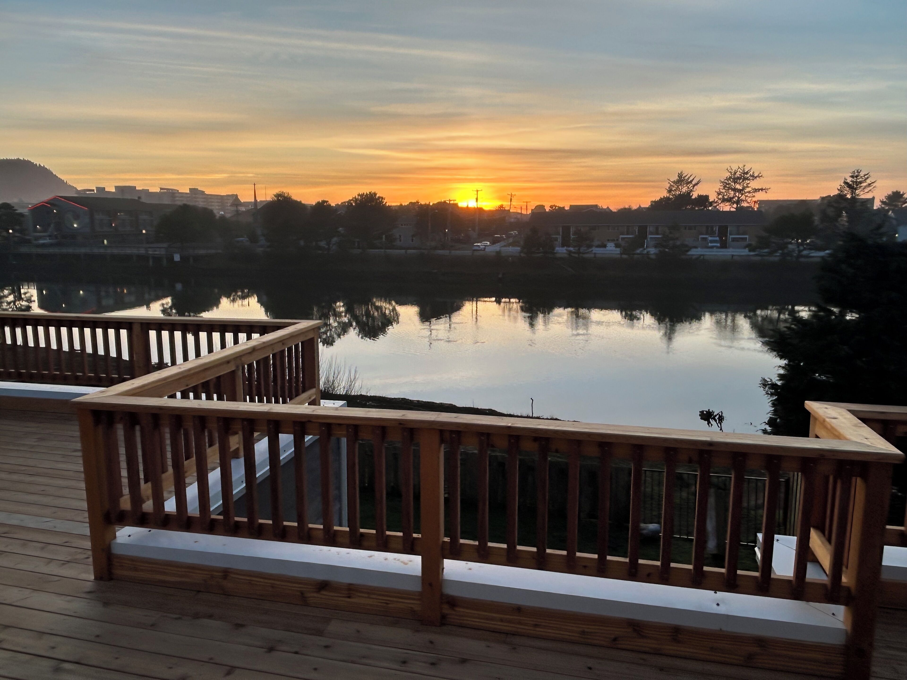 Une terrasse en bois, dotée de rampes de protection, qui offre une vue sur un lac paisible au coucher du soleil.