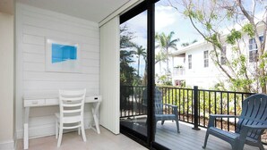 A balcony with a white desk and two chairs, a glass door leading outside, and a view of a white building with palm trees. - Captain's Haven Sanibel, with Pergola on East End (Sanibel)