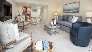 A spacious living room with a grey sofa, a blue armchair, and a wooden chair. There is a dining area with a table and chairs, and a kitchen in the background. - Captain's Haven Sanibel, with Pergola on East End (Sanibel)