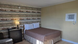 Een slaapkamer met een houten wandpaneel, een bed met een bruine plaid, een nachtkastje met een lamp en een wandairconditioner.