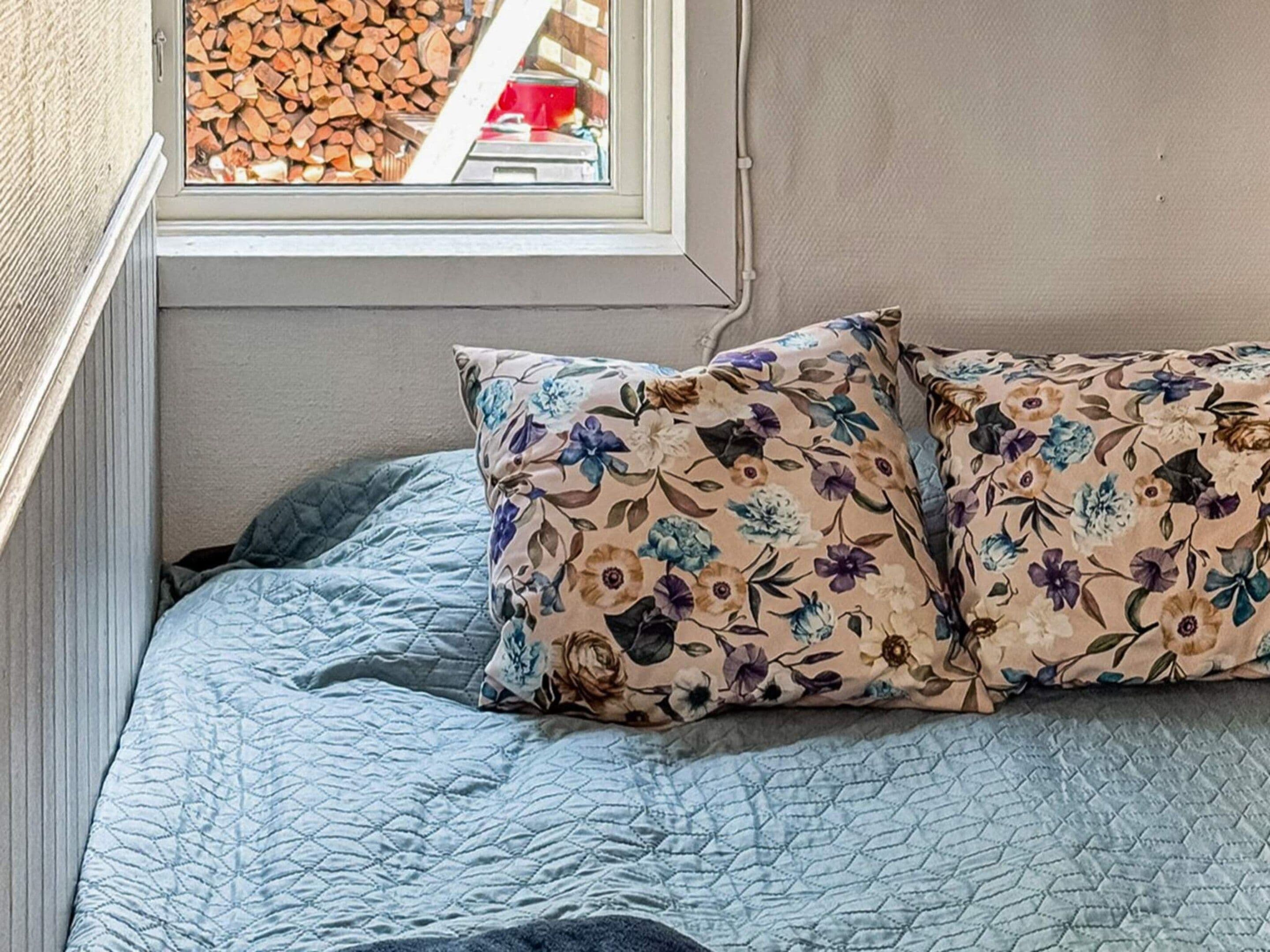 Una cama con un cojín floral y una colcha azul acolchada, una ventana con vistas a leña apilada y una pared con textura.