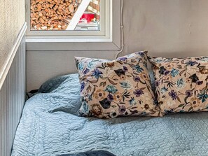 A bed with a floral pillow and a quilted blue blanket, a window with a view of stacked firewood, and a textured wall. - 7 Person Holiday Home in Hestra (Hestra)
